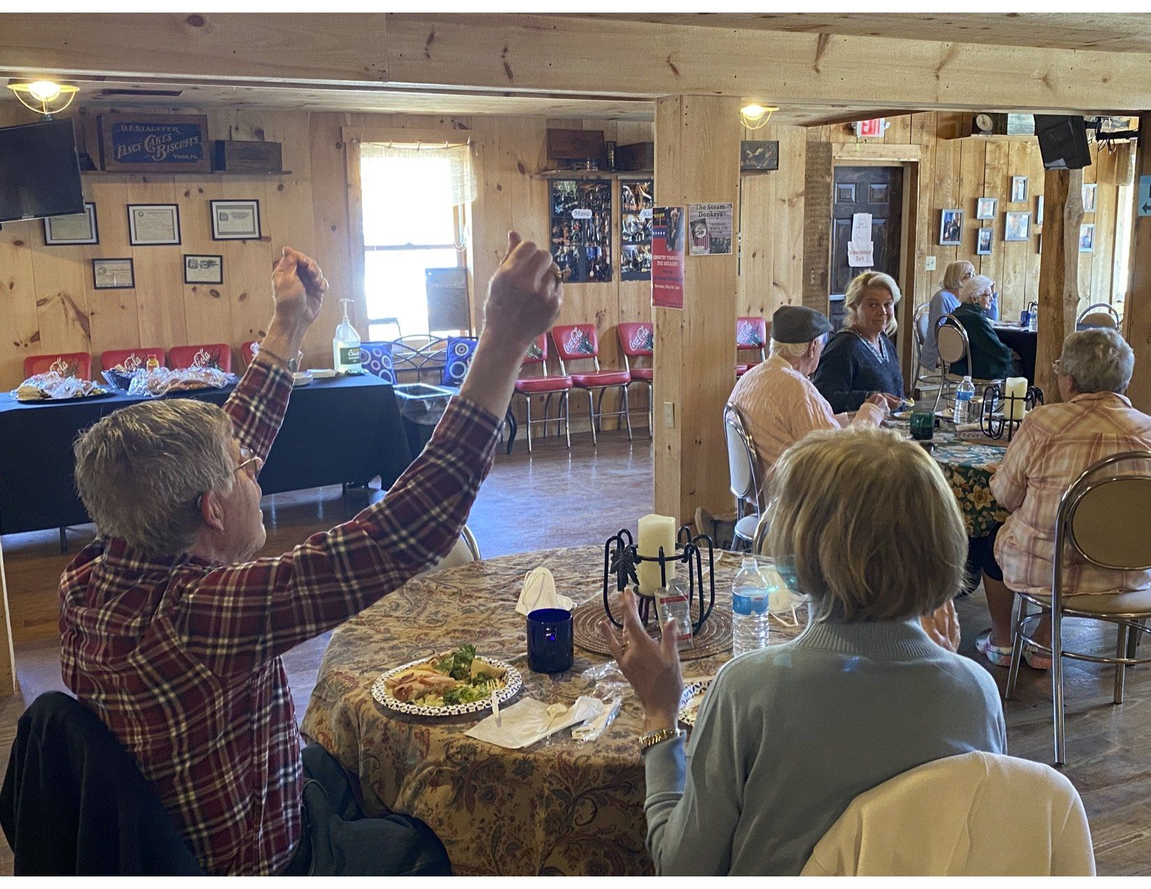 People at tables in a wooden-paneled room. One man raises arms. Some dine, while others look on.