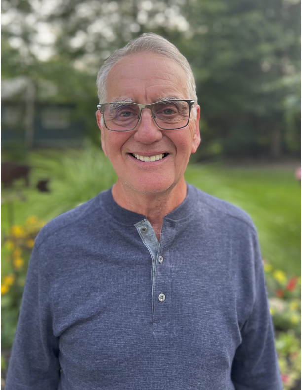 Man with glasses smiles outdoors, wearing a blue Henley shirt.