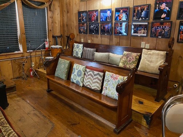 Wooden church pew with pillows, photos on wood paneled wall. Stage with drums in background.