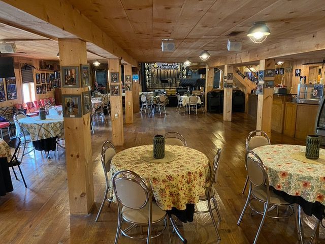 Interior view of a restaurant with round tables, chairs, and wooden beams. Tables have floral tablecloths.