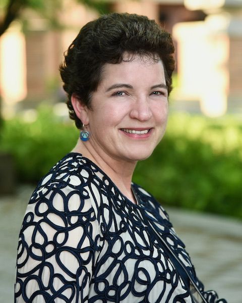 Woman in a navy and white patterned top smiles outdoors. Short dark curly hair, blue earrings.