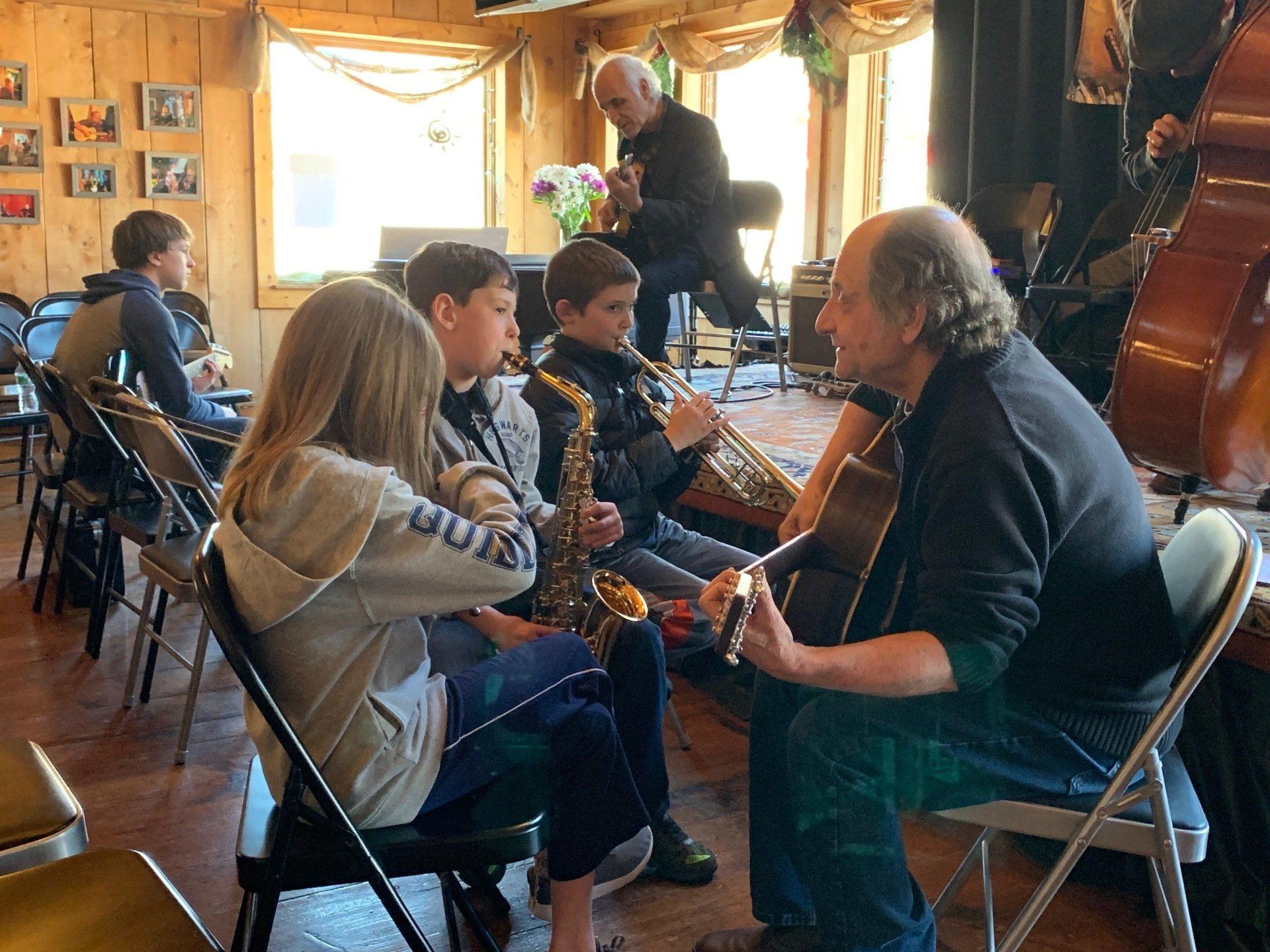 Musicians with instruments, teaching students. Indoors with natural light, stage, and chairs.