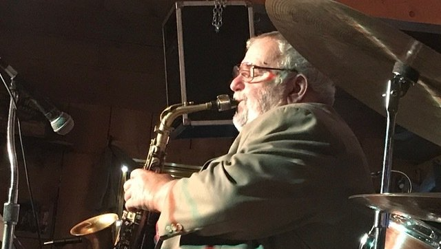 Saxophonist playing, stage lit, dark background, saxophone, microphone, cymbals, glasses, jacket.