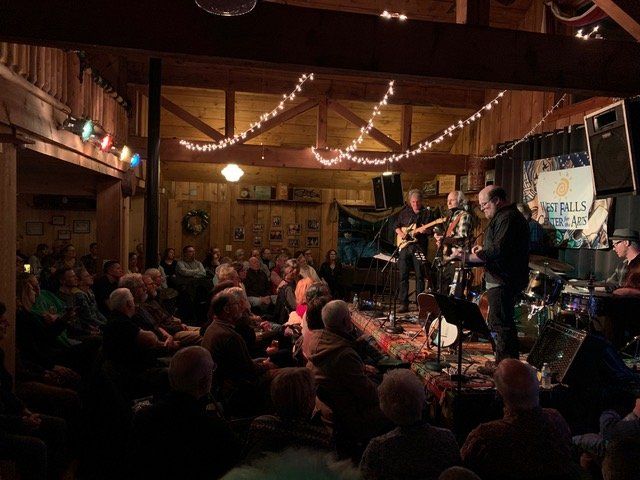 Band performing on stage in a dimly lit venue, playing to a seated audience. Overhead string lights.