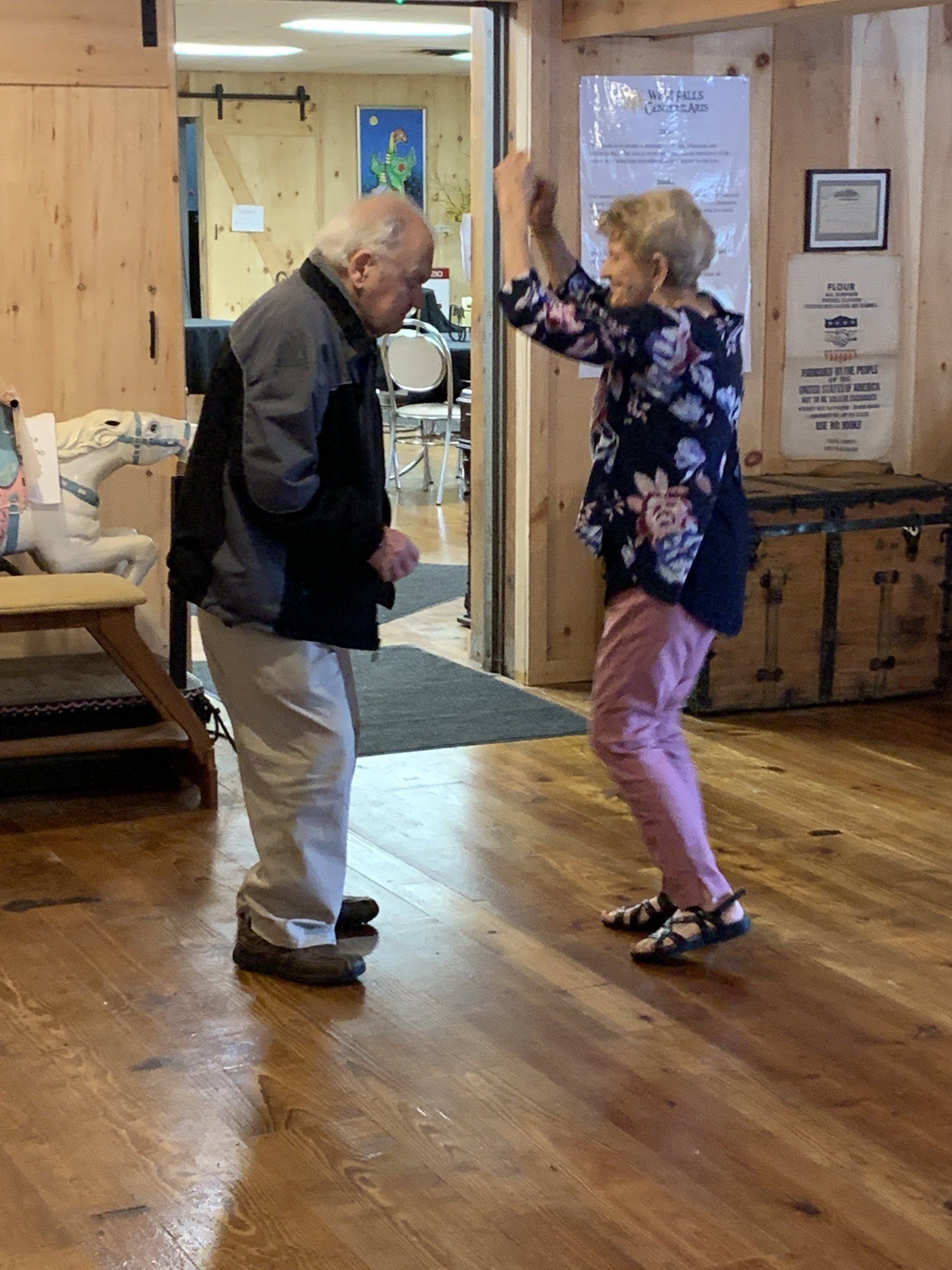 Elderly couple dancing on a wood floor in a building. The woman has her hands raised, and they are both smiling.