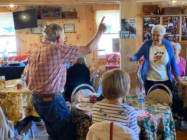 Man pointing and woman dancing in a room with tables and other people seated.