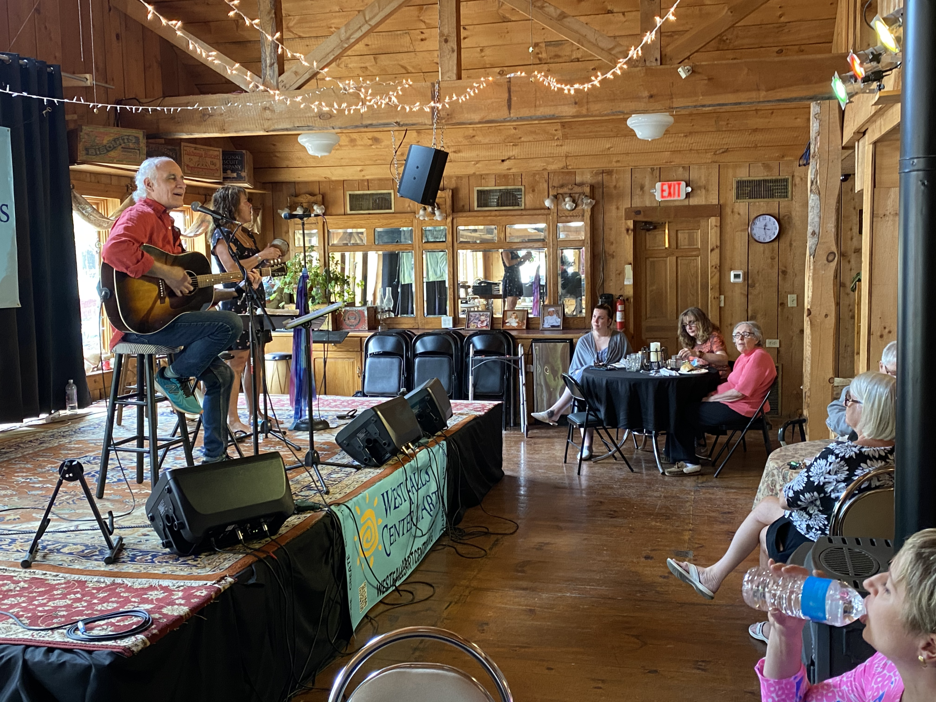 Musicians performing on a stage in a wooden-paneled room, with audience members seated at tables.