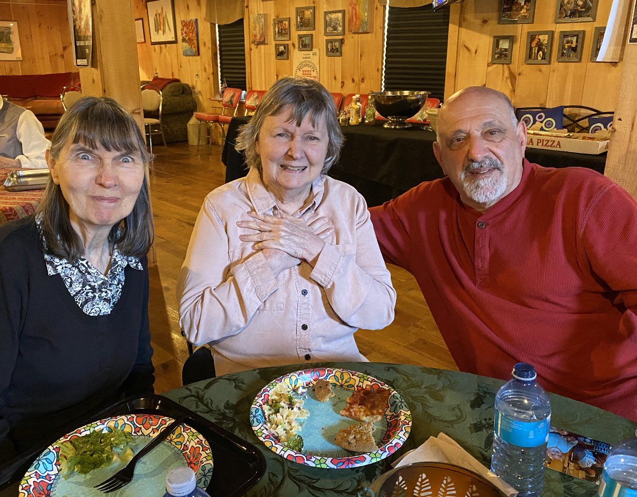 Three people smiling at a table with plates of food. Middle person with hands on chest. Interior setting.