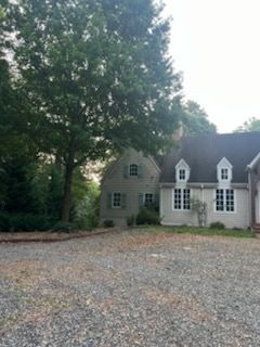 Gravel driveway beside a gray house and tall trees in soft evening light