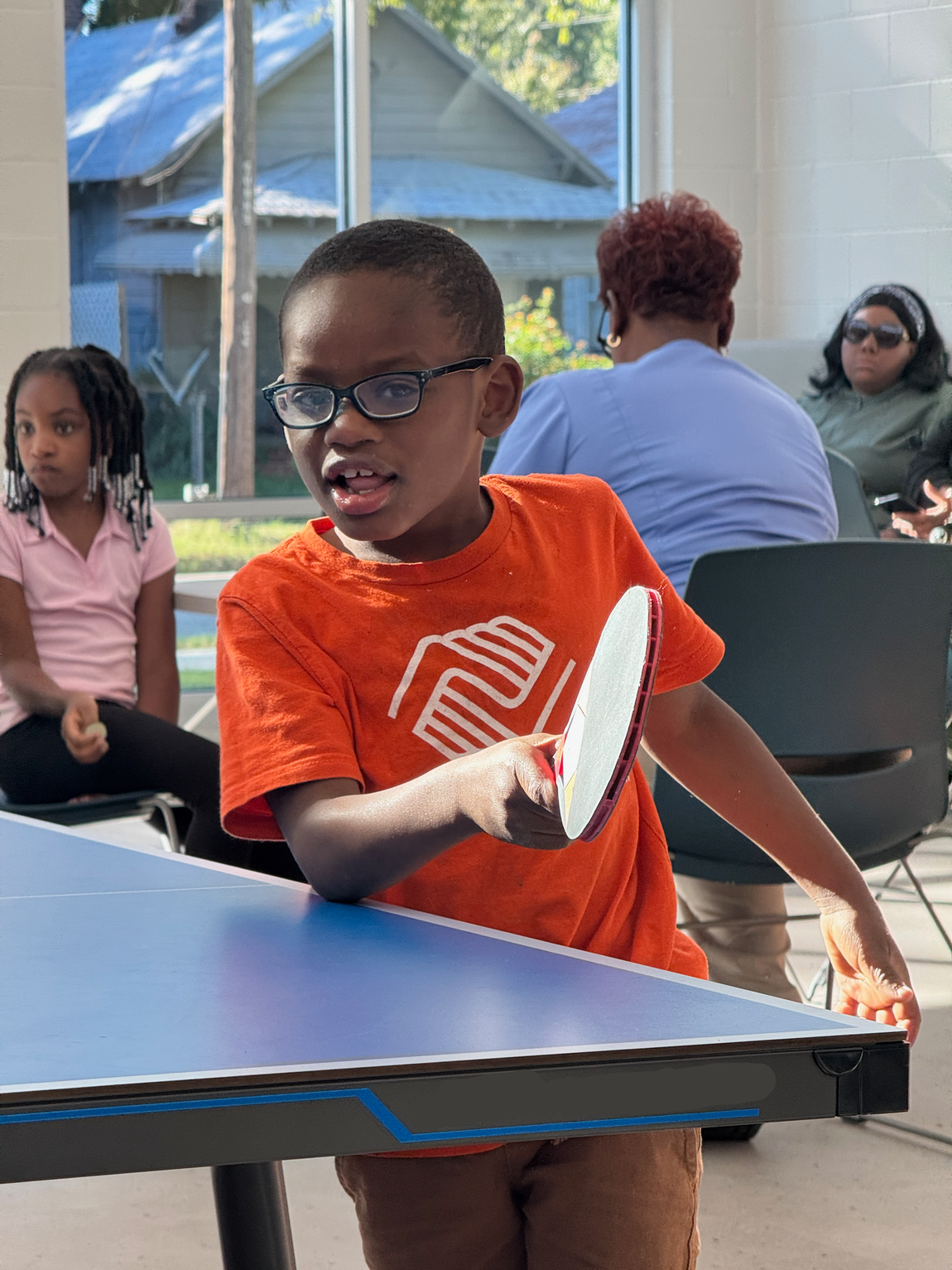 Child in orange shirt playing table tennis indoors, holding a paddle at the table.