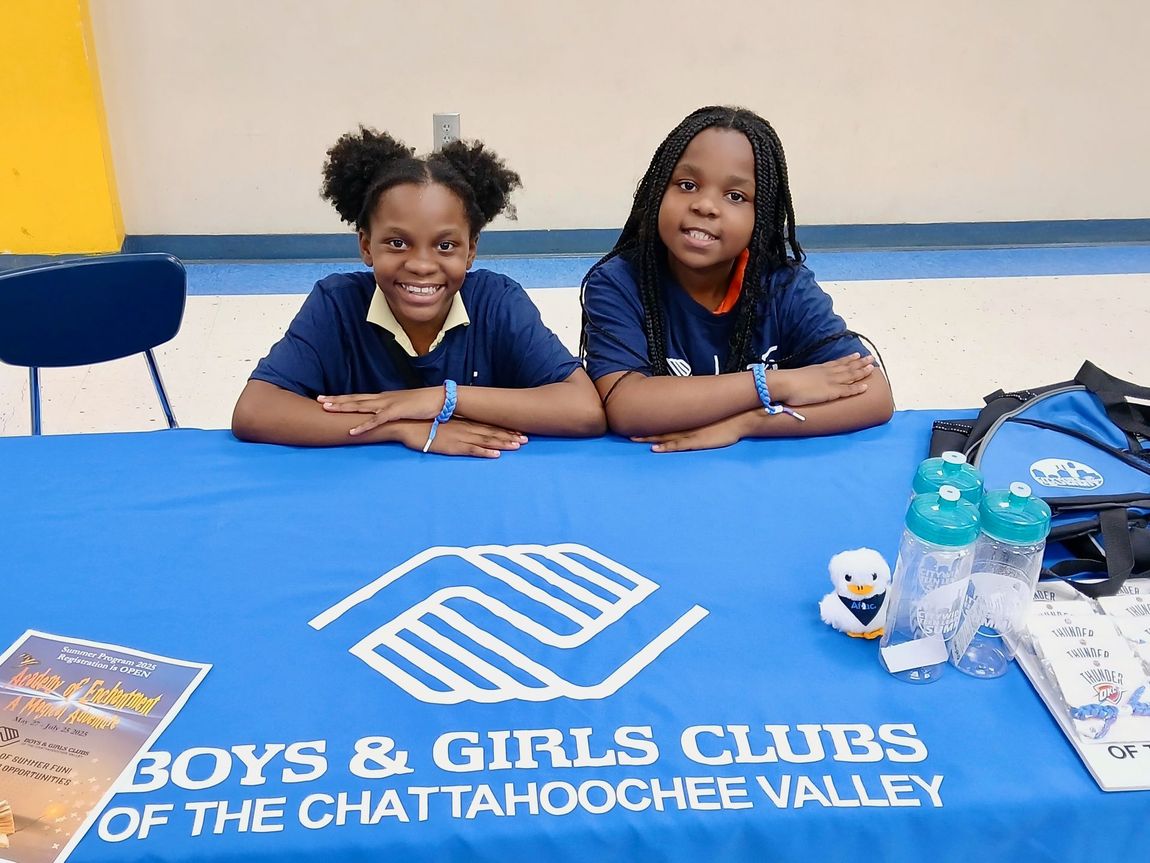 Two smiling children at a Boys & Girls Clubs table with flyers and bottles in a gym-like room