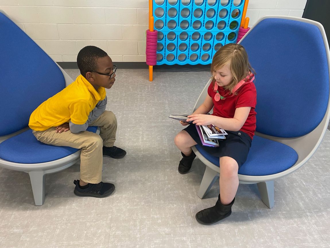 Two children sitting on blue chairs, talking in a playroom with a colorful wall game.