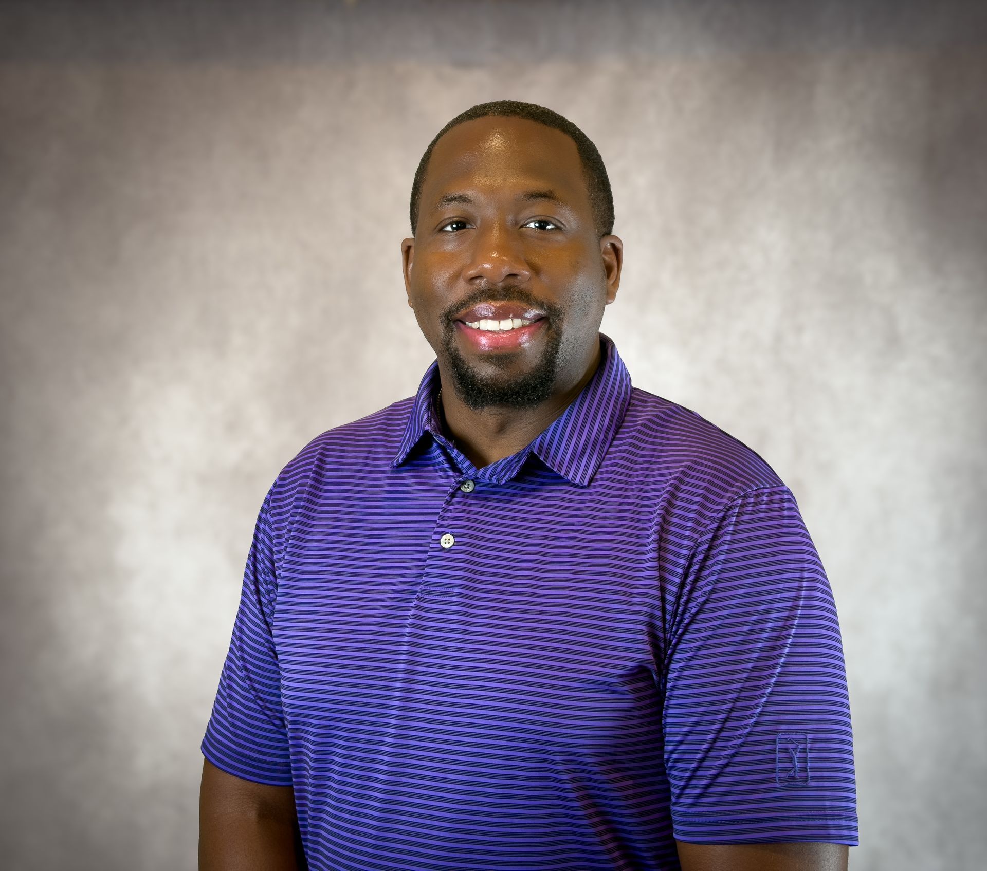 Smiling man in a purple striped polo shirt against a gray studio backdrop