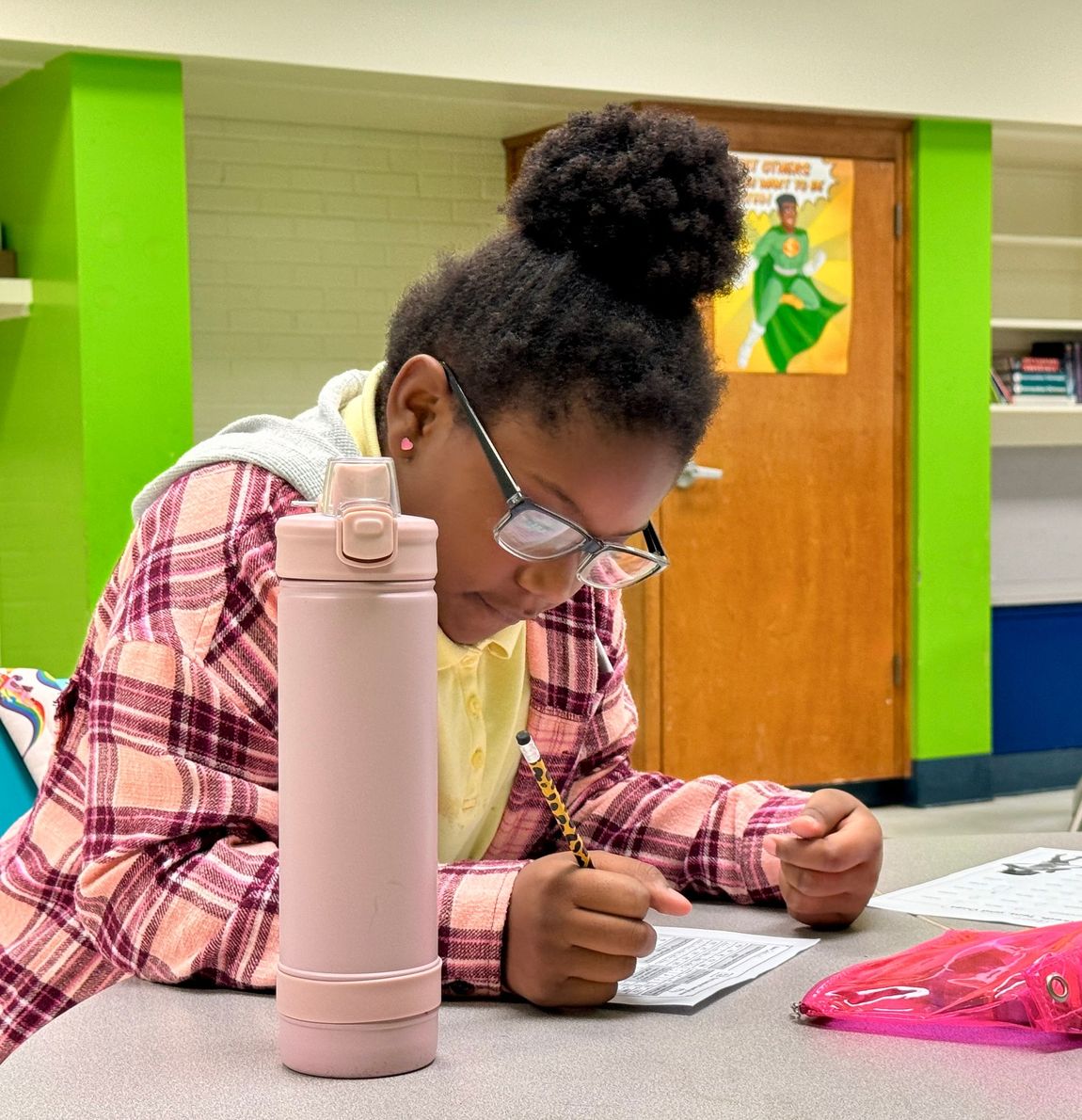 Student writing at a table in a classroom, with a pink water bottle in the foreground.