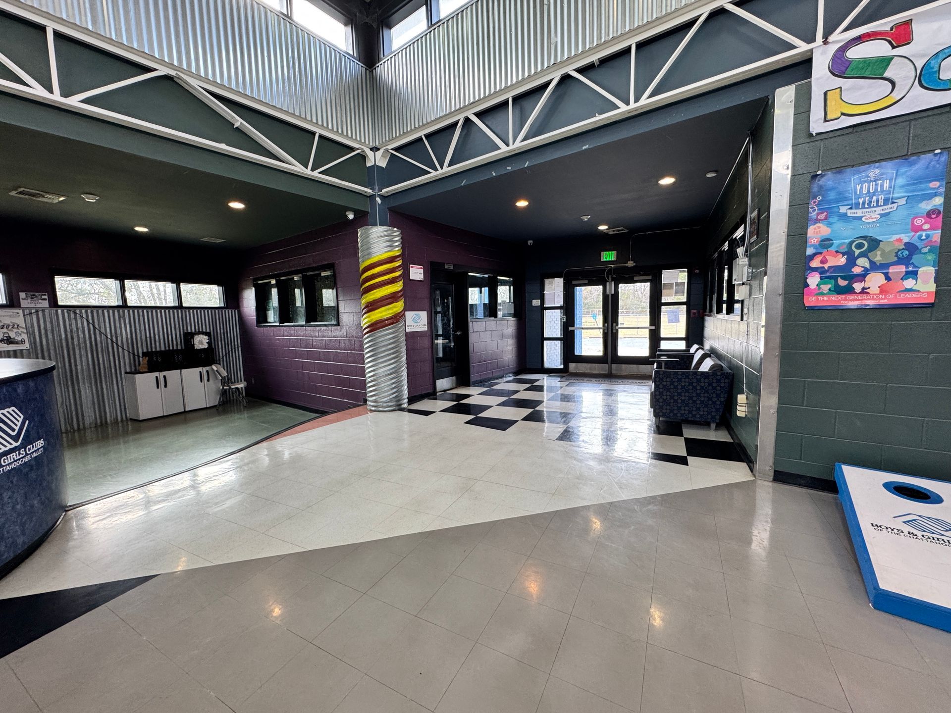 Empty indoor lobby with checkered tile floor, gray walls, and a yellow-black striped pillar near glass doors.