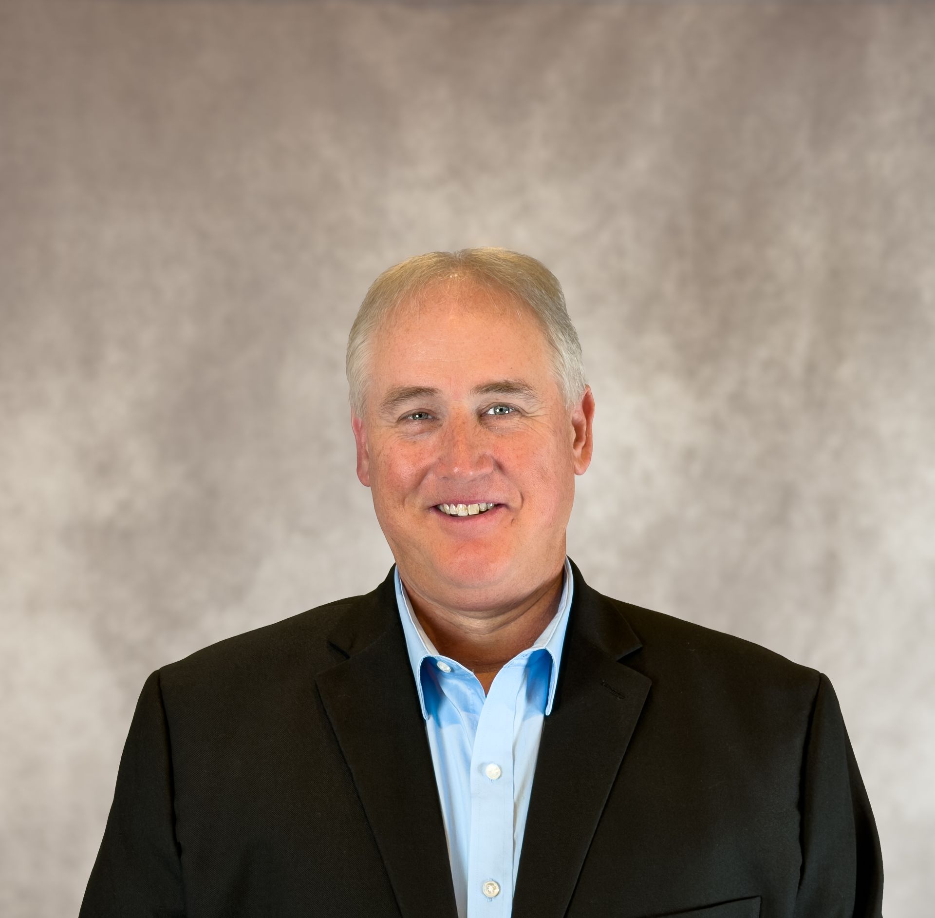 Smiling man in a black blazer and blue shirt against a gray studio backdrop