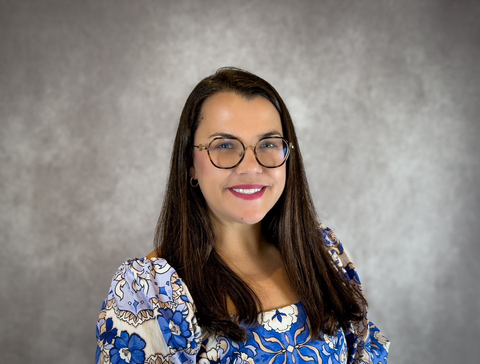 Smiling woman with glasses in a blue floral top against a gray studio backdrop