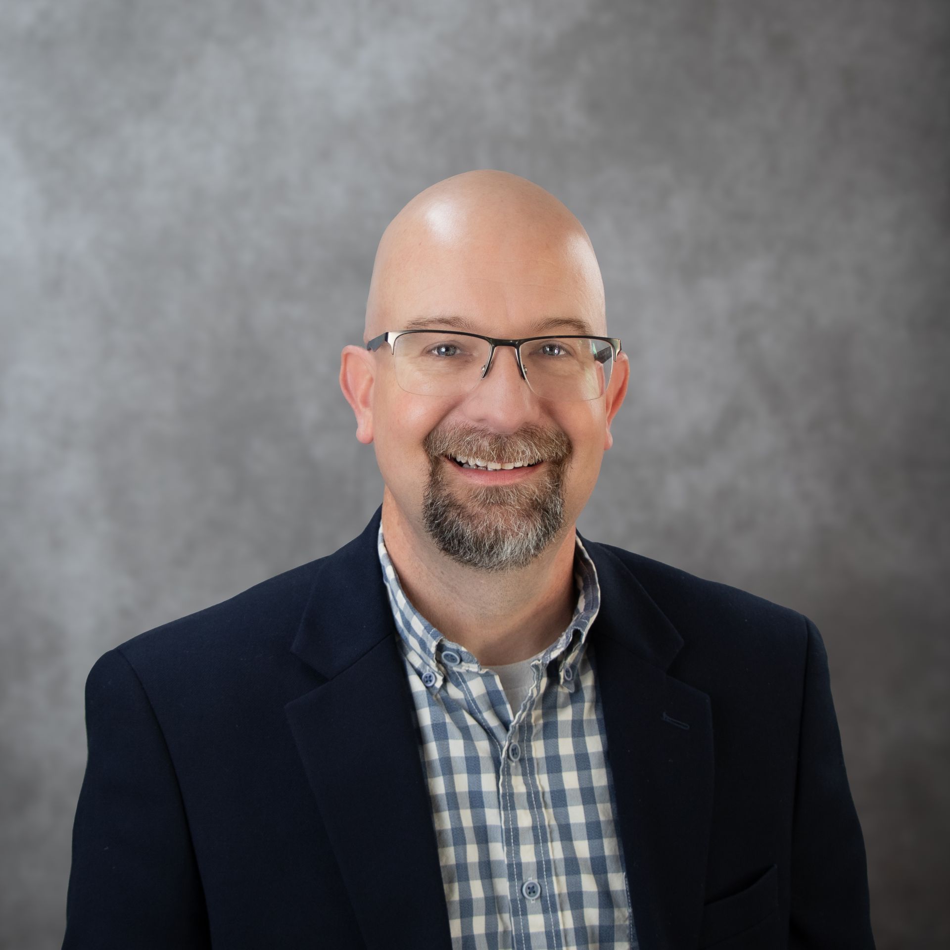 Smiling bald man in glasses wearing a dark blazer and plaid shirt against a gray backdrop
