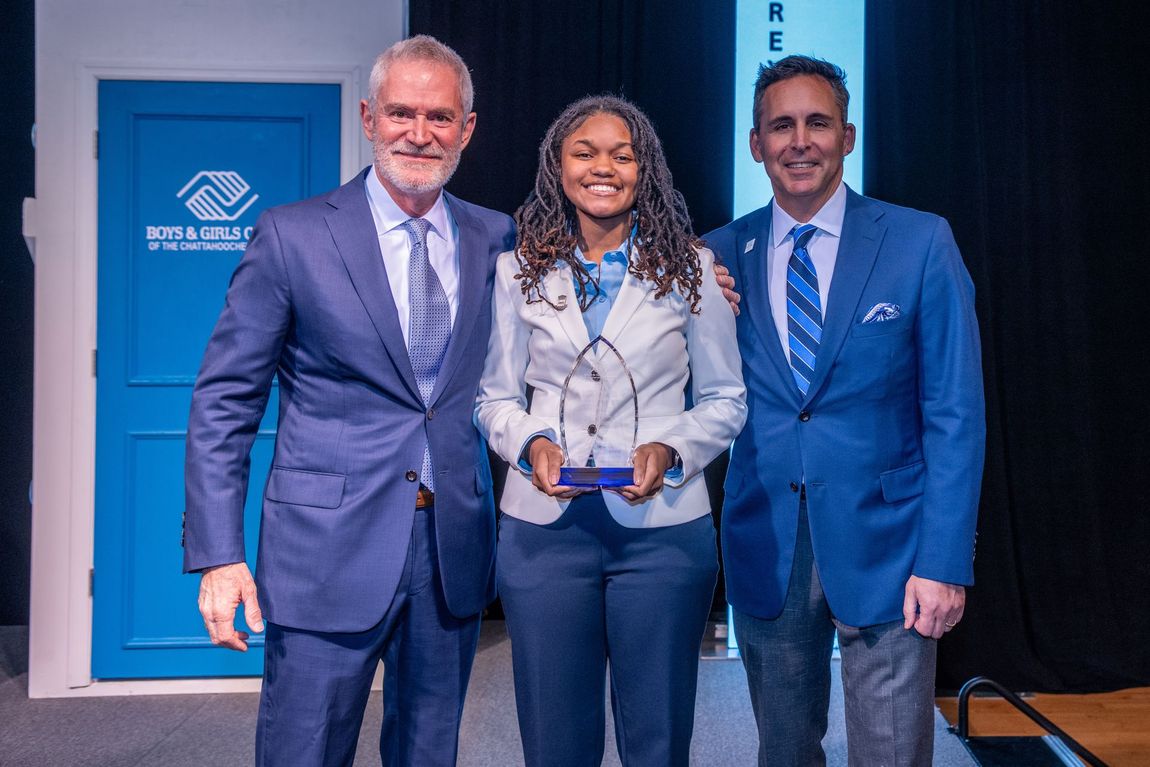 Three people in blue suits posing with a plaque at a ribbon-cutting event indoors