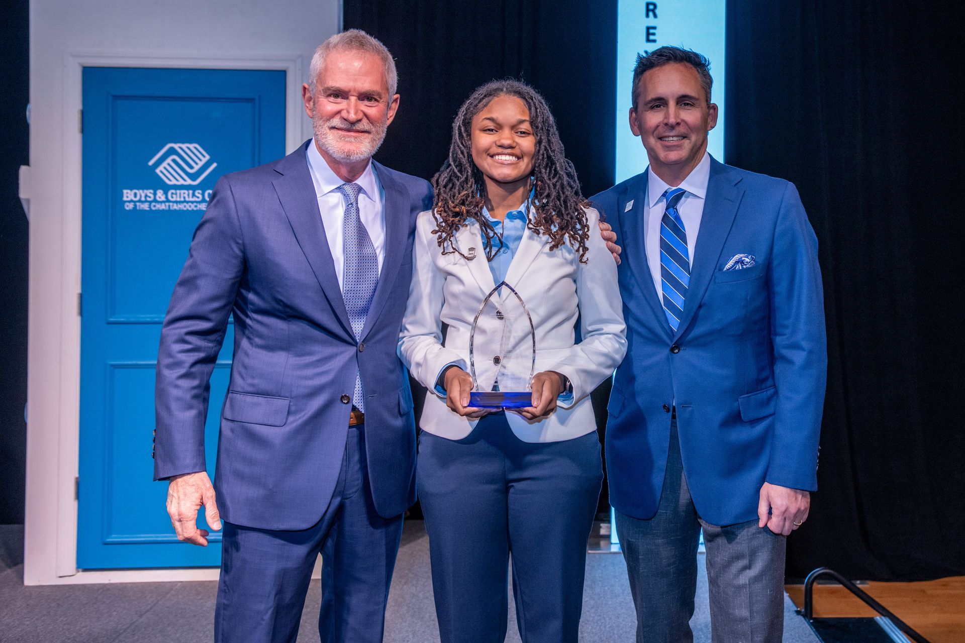 Three people in blue suits posing with a plaque at a ribbon-cutting event indoors