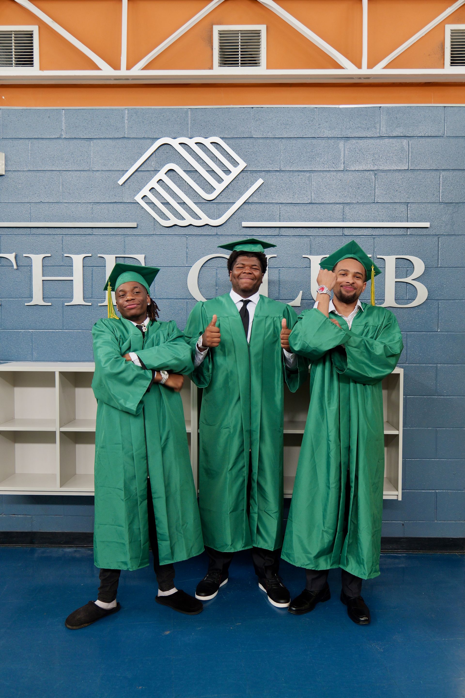 Three graduates in green gowns and caps posing in front of a Boys & Girls Club wall