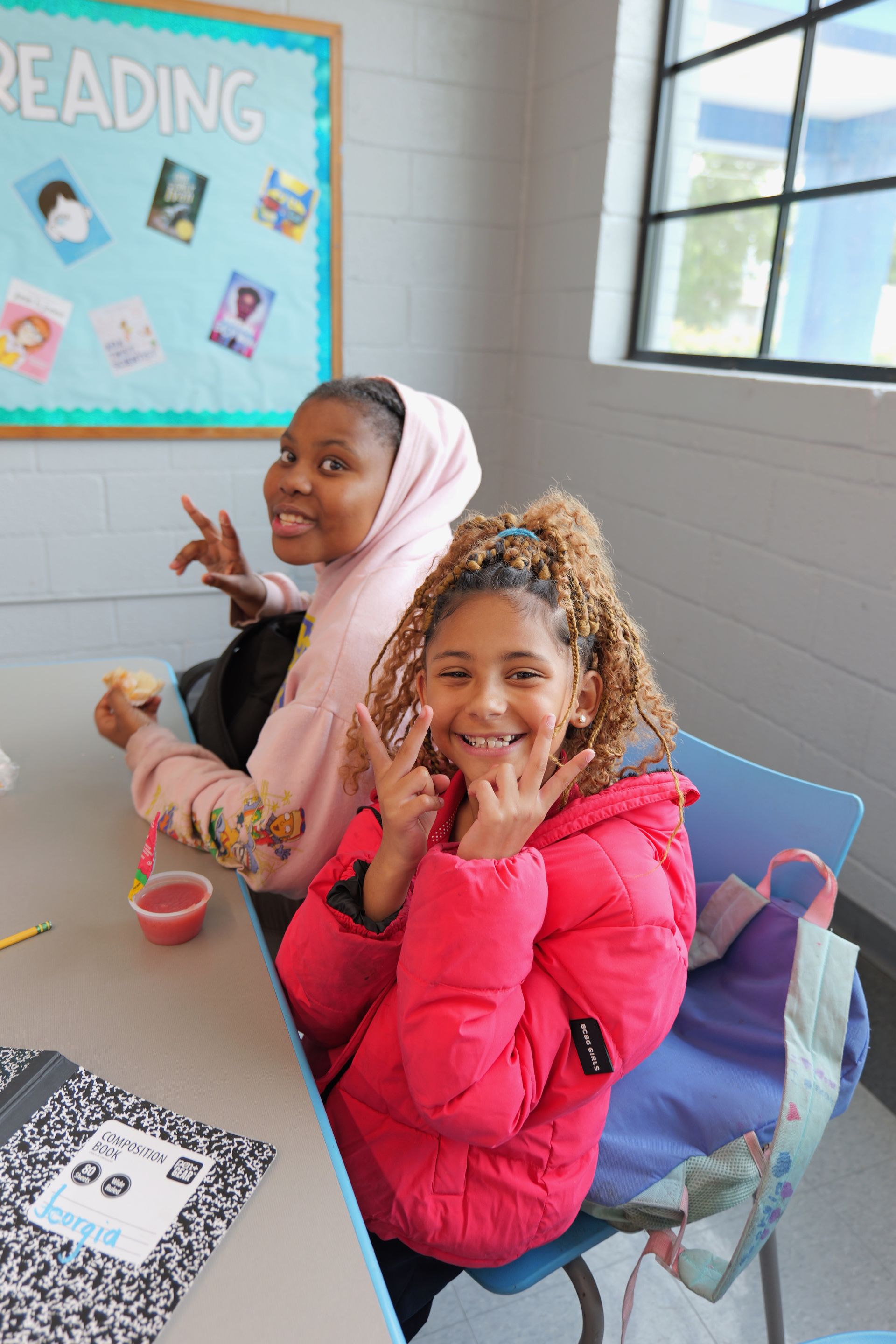 Two children smiling at a table in a bright classroom, with a reading bulletin board behind them.