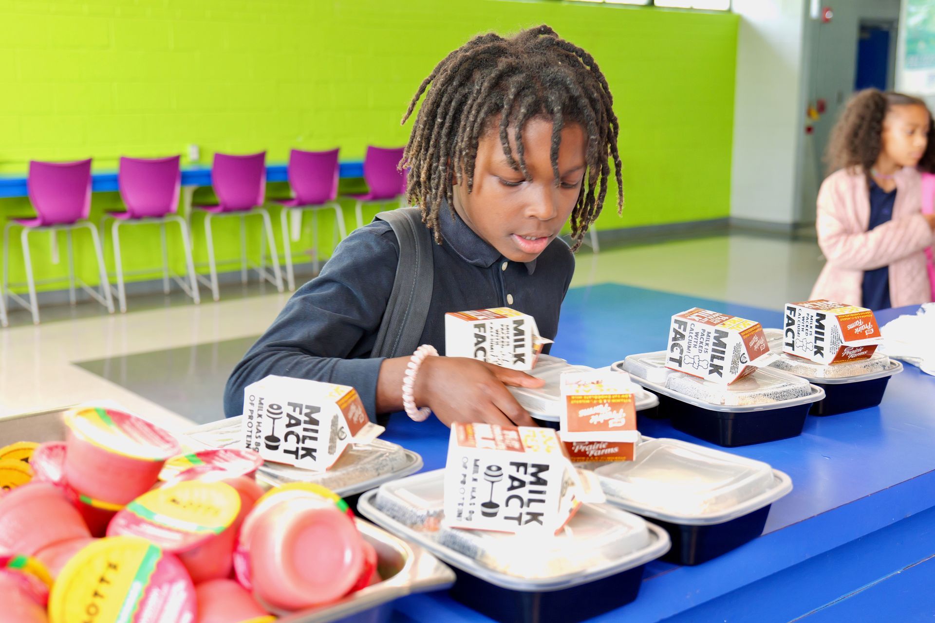 Child eating lunch at a blue cafeteria table, with trays, milk cartons, and colorful plates nearby.