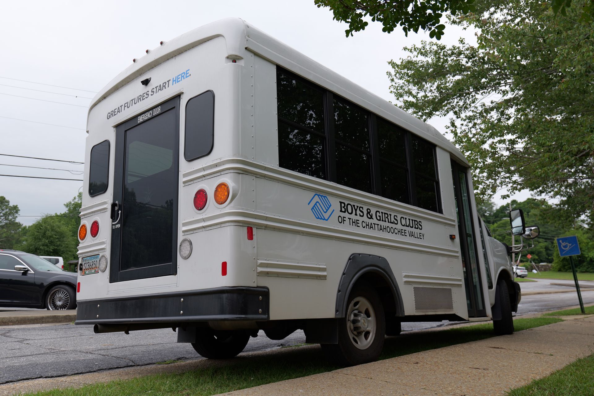White shuttle bus parked on a tree-lined street beside a sidewalk and parked car
