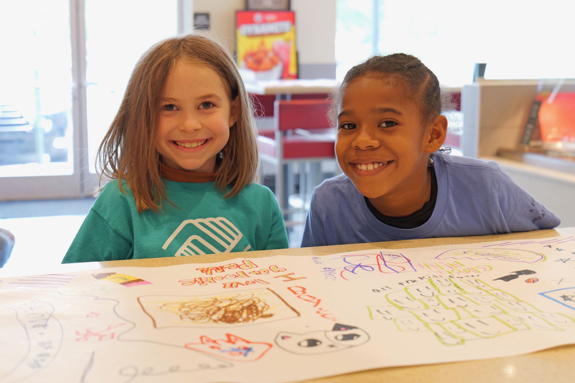 Two smiling children at a table with colorful drawings on paper in a classroom.