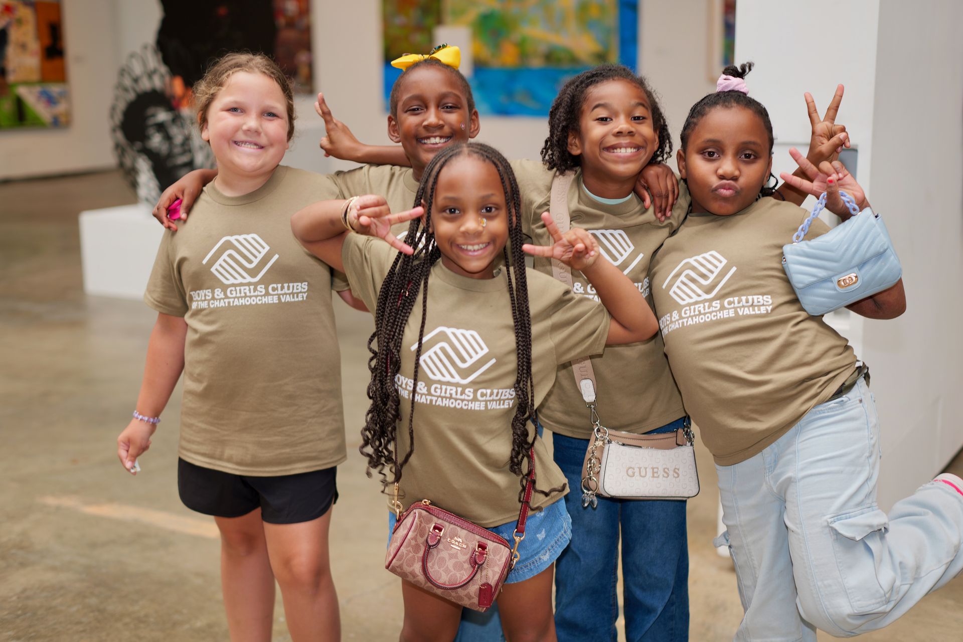 Five children posing and smiling in a hallway, wearing matching tan shirts