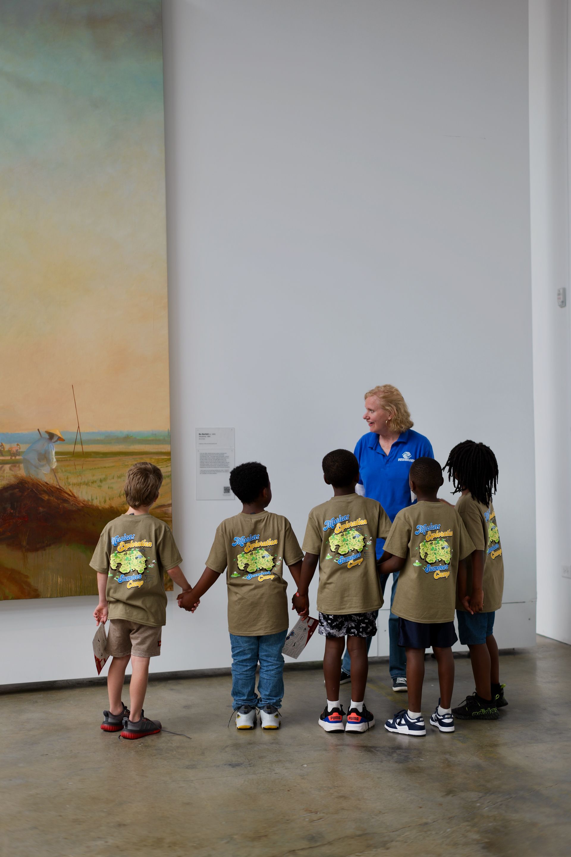 Five children standing in a museum, facing a woman in blue near a large wall mural.