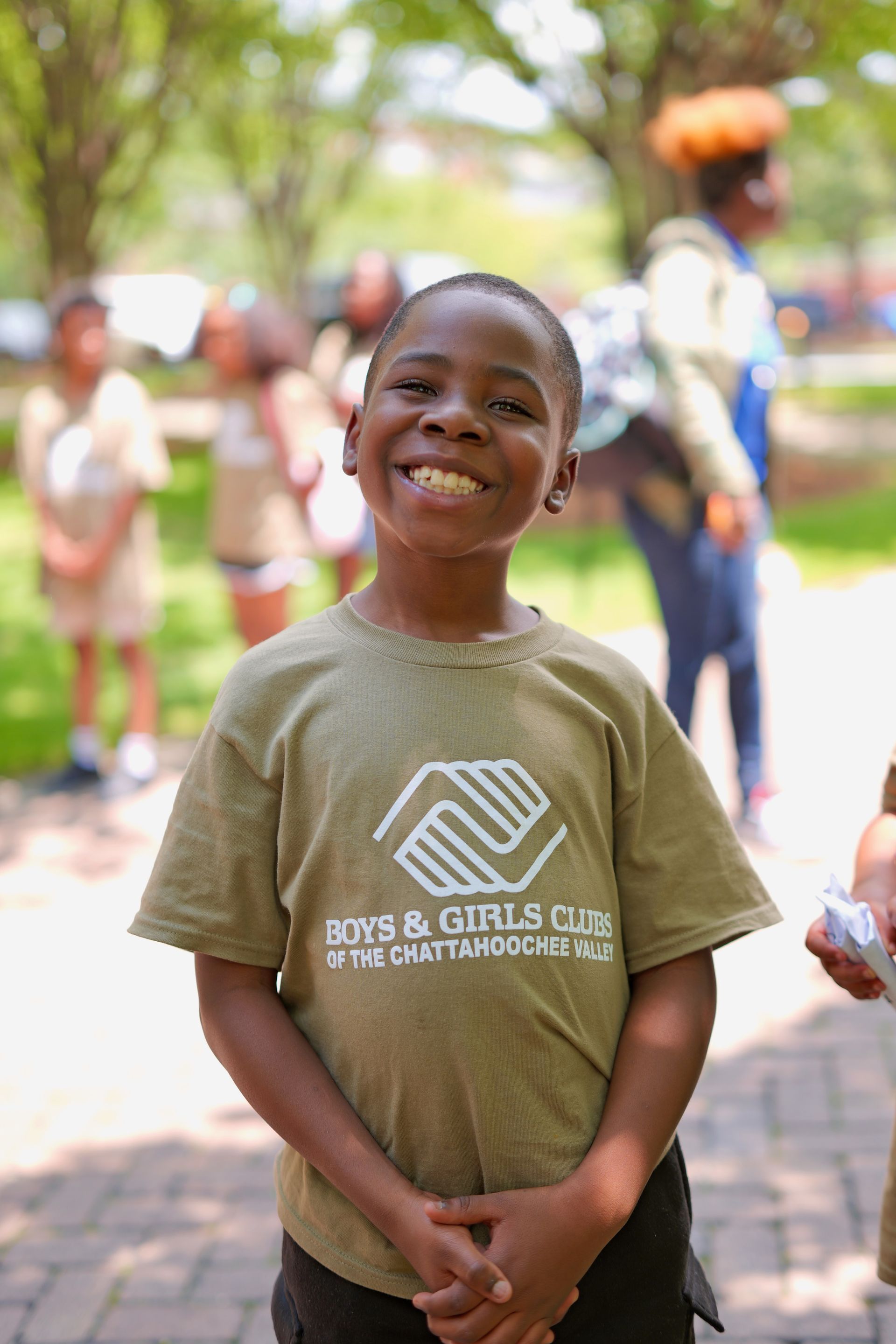 Smiling child standing outdoors in a tan Boys & Girls Club T-shirt, with people blurred in the background