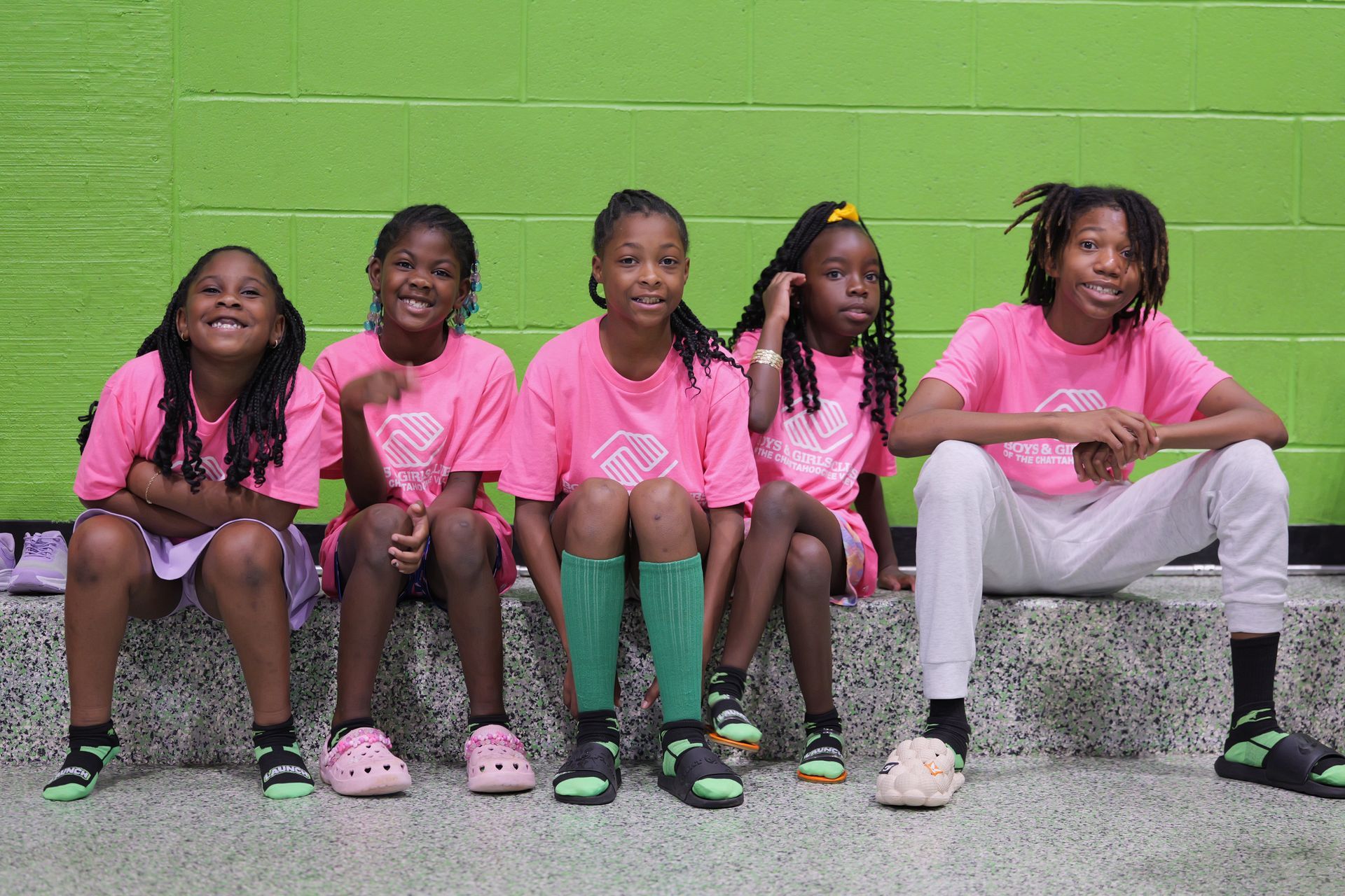 Five children in pink shirts sit on a bench in front of a green corrugated wall.