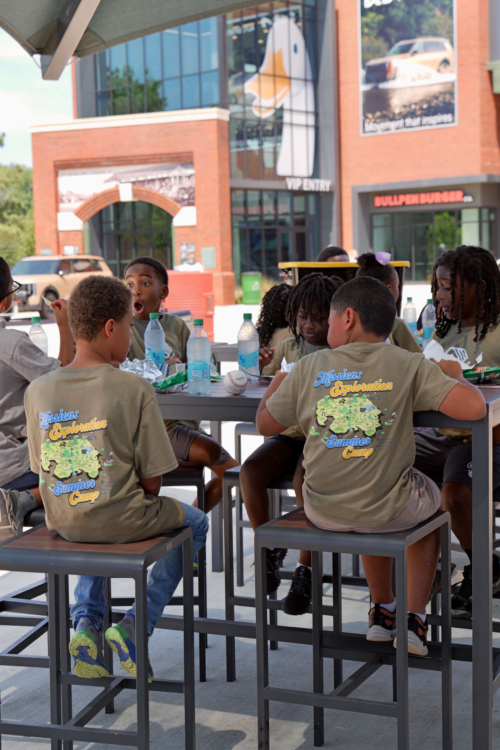 People seated at tall tables outside a brick building with glass windows and posters.