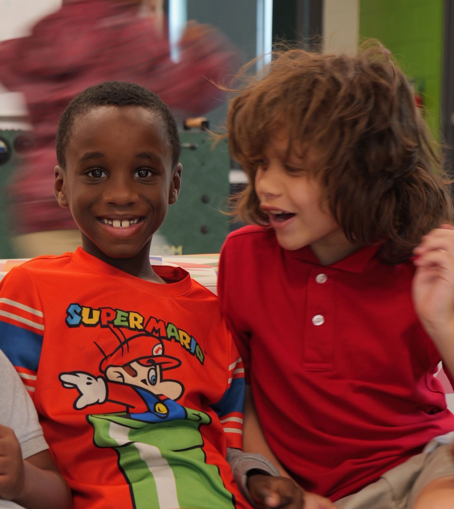 Two children smiling on a couch, one in a red Super Mario shirt and the other in a red polo.