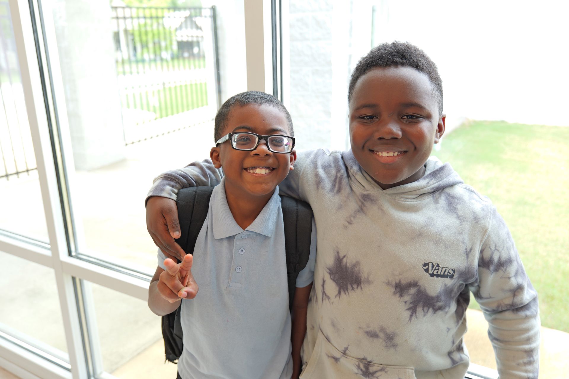 Two children smiling indoors, one with an arm around the other, near a bright window.