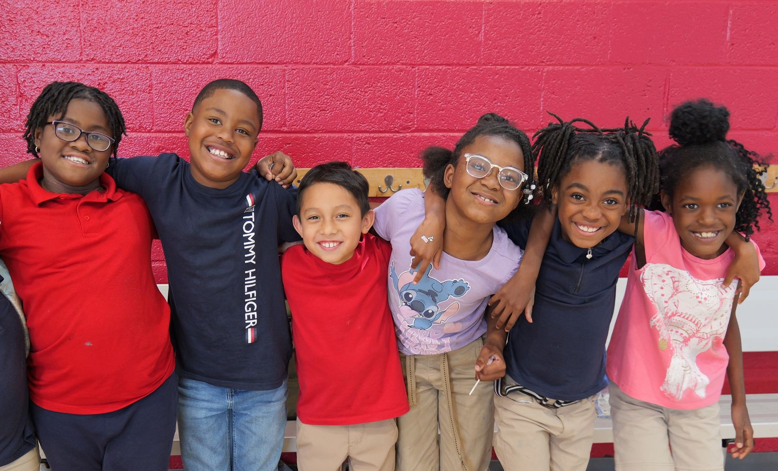 Six children stand smiling shoulder to shoulder against a red wall, wearing red, navy, and pink shirts.
