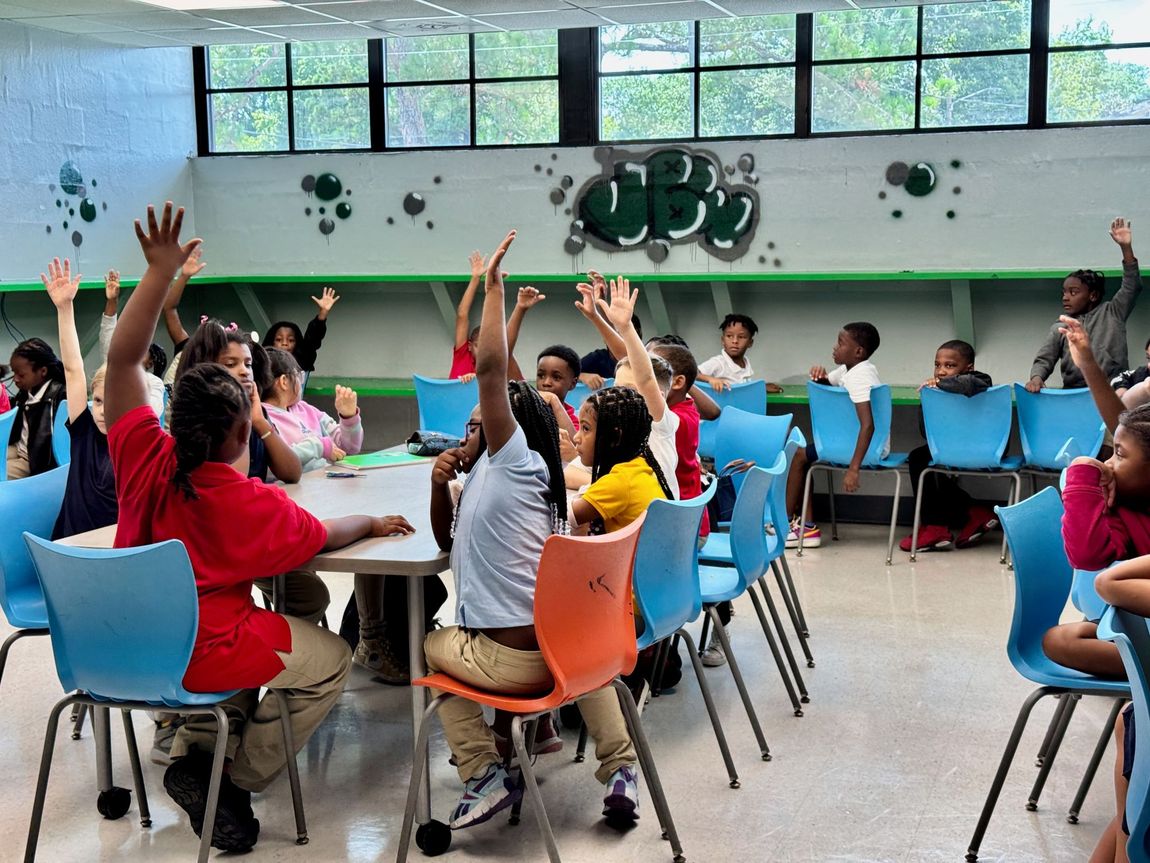 People seated in a classroom raise hands during a group lesson with blue chairs and large windows.