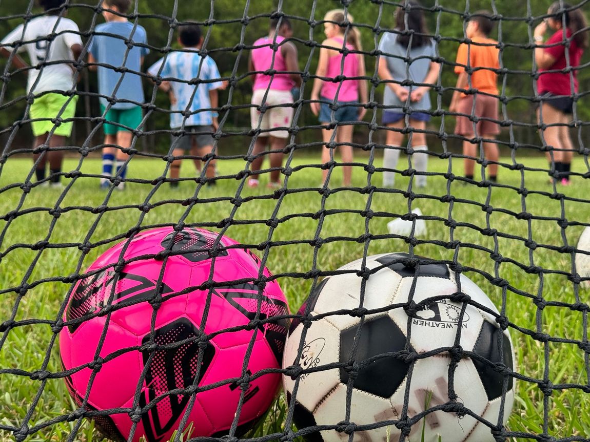 Two soccer balls by a goal net, with children standing on the field in the background.