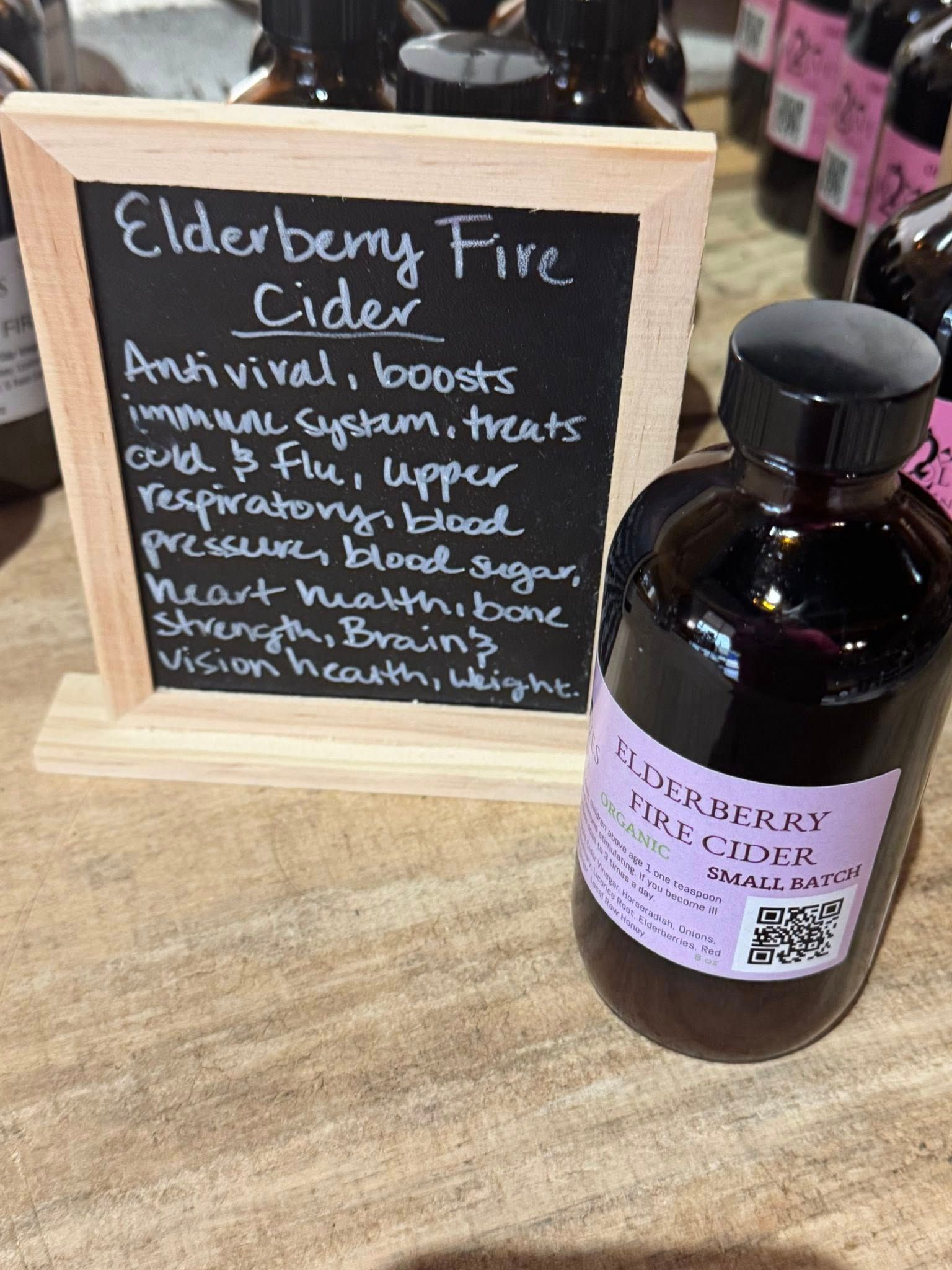 Elderberry Fire Cider sign beside a dark glass bottle with a purple label on a wooden counter