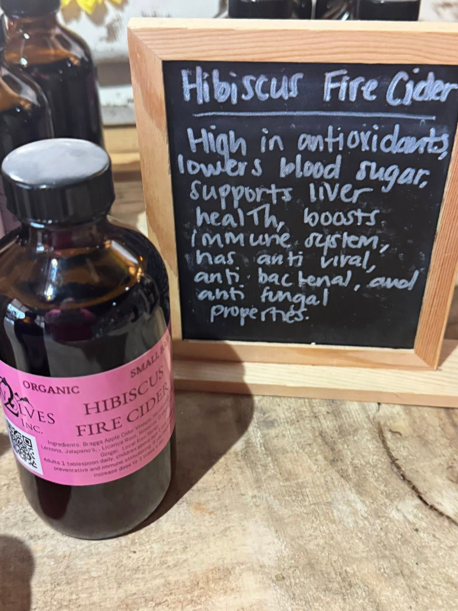 Brown glass cider bottle beside a chalkboard sign advertising hibiscus fire cider with local honey and herbs