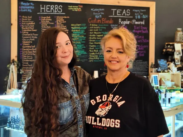 Two women standing in a tea shop, with a chalkboard menu behind them and smiling at the camera.