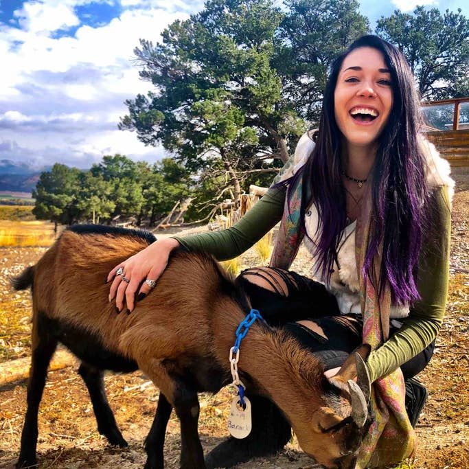 Woman smiling and petting a brown goat in an outdoor setting with a mountain backdrop.