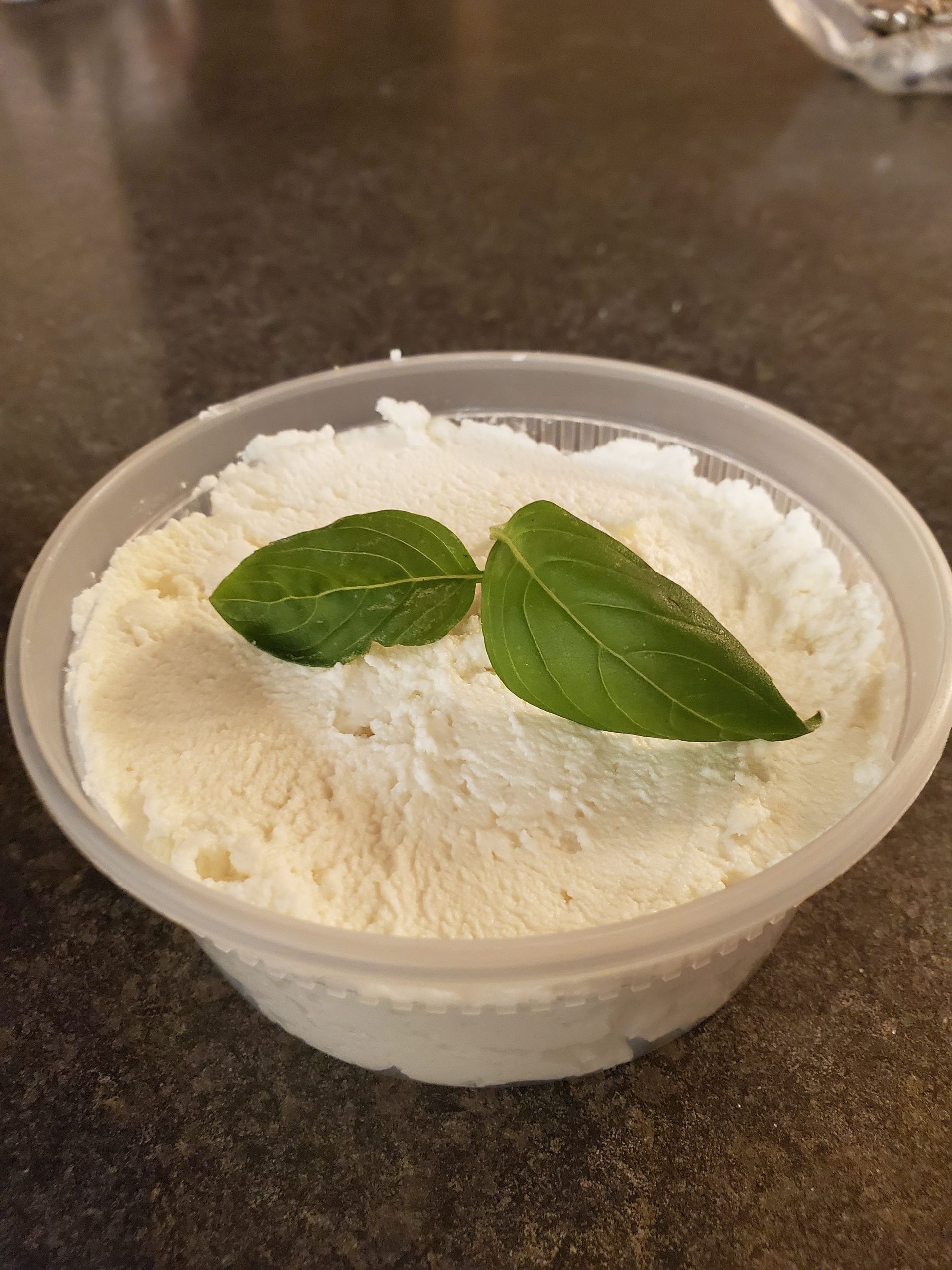 White, creamy food in a clear plastic container, garnished with two green basil leaves on a dark surface.