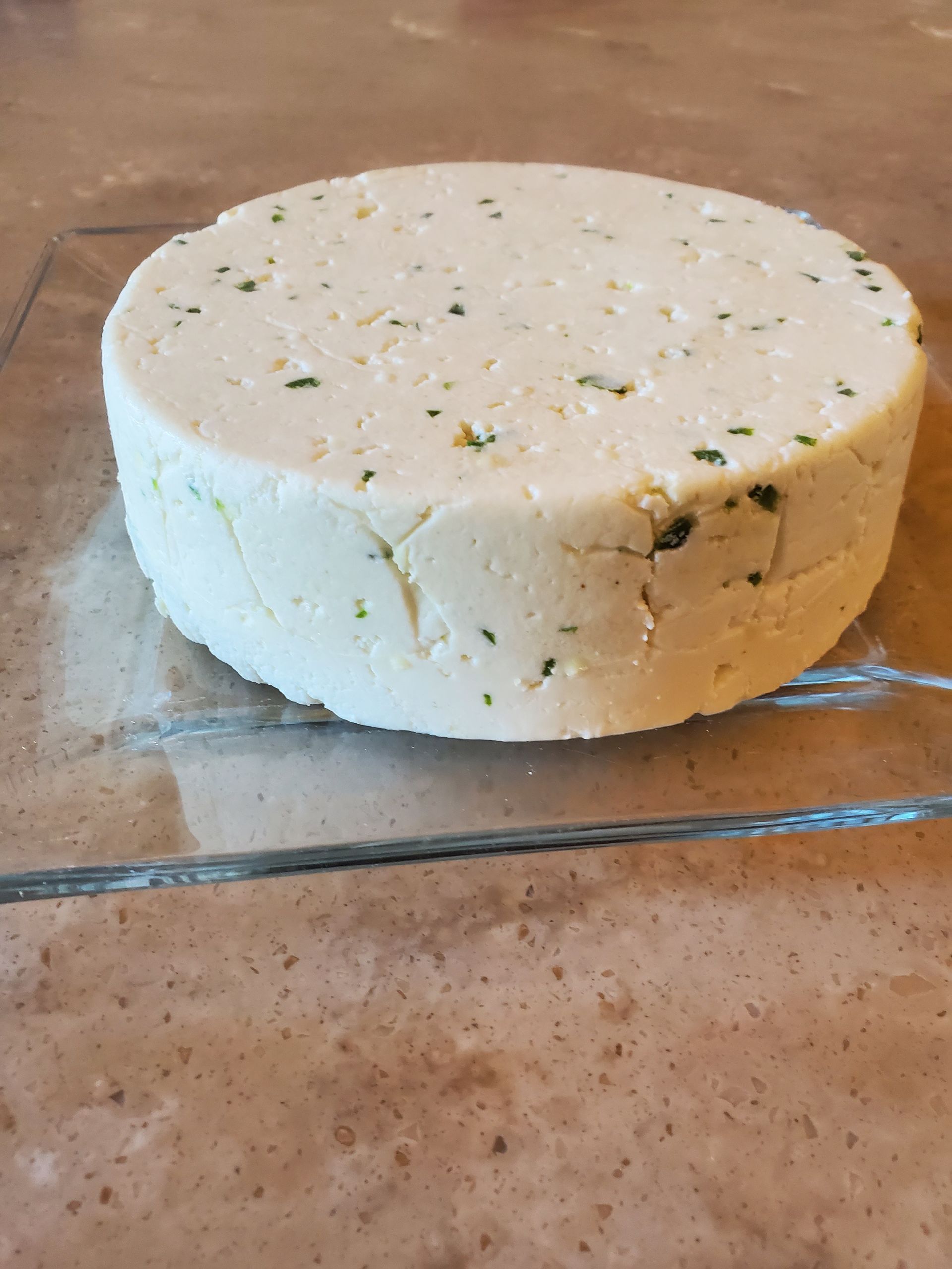 Round cheese, off-white with green flecks, on a clear glass plate, set on a brown surface.