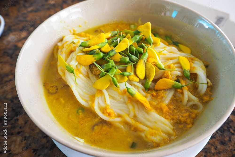 Bowl of yellow noodles in broth, topped with yellow flowers and green buds.