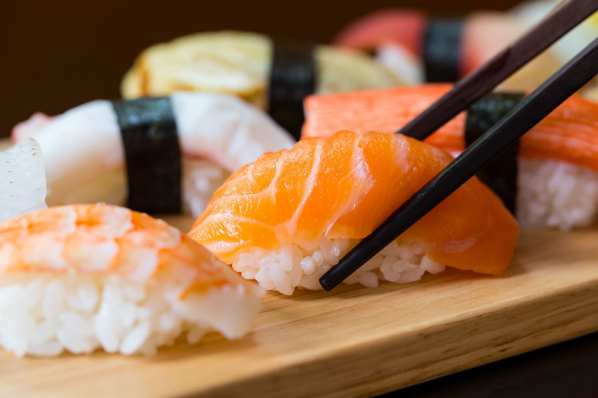 Sushi nigiri on a wooden board; chopsticks are picking up a salmon piece.