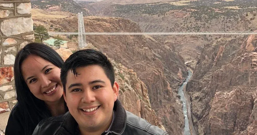 Couple smiling in front of Royal Gorge Bridge; mountainous canyon in background.