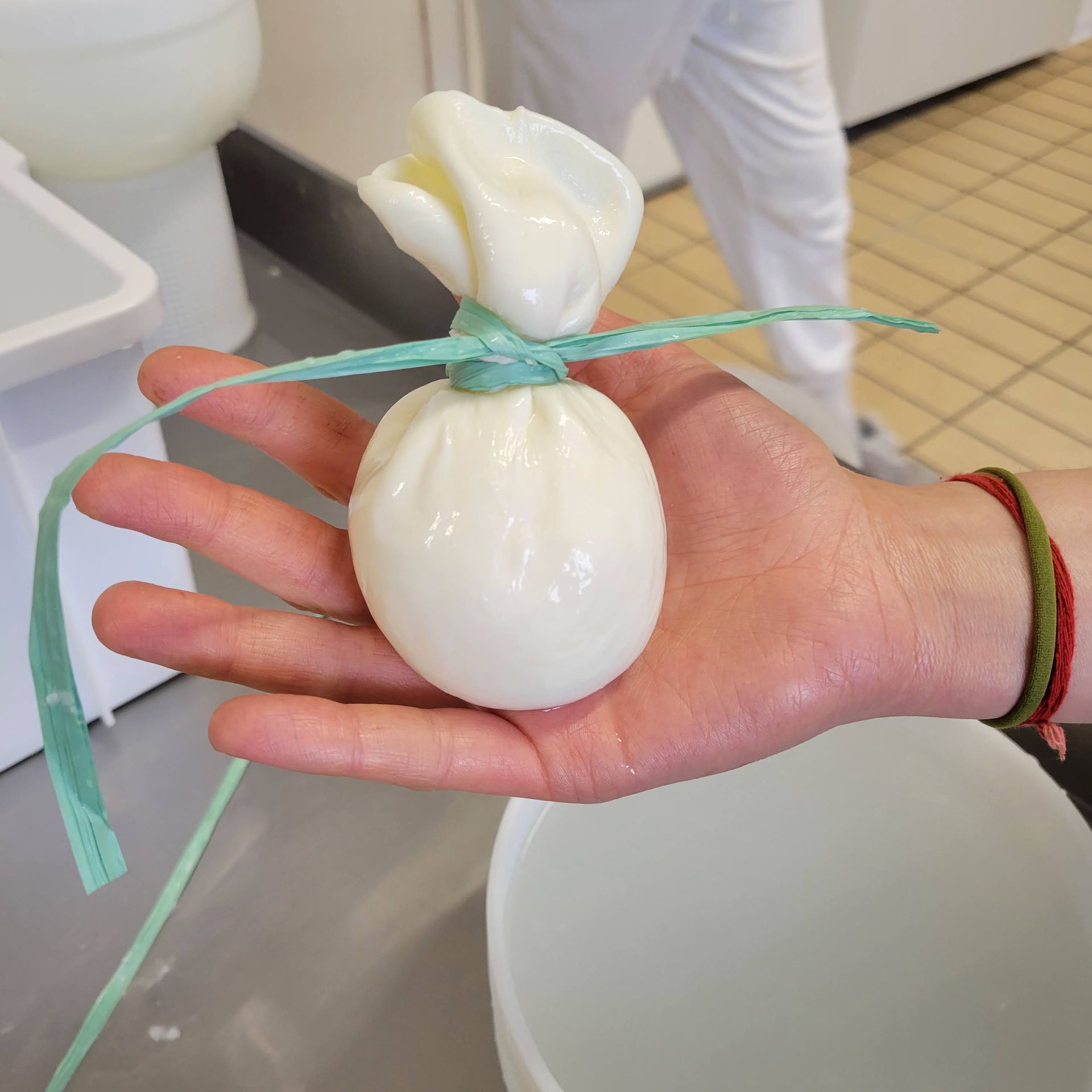 A hand holding a tied-up ball of white cheese, likely burrata, in a kitchen setting.