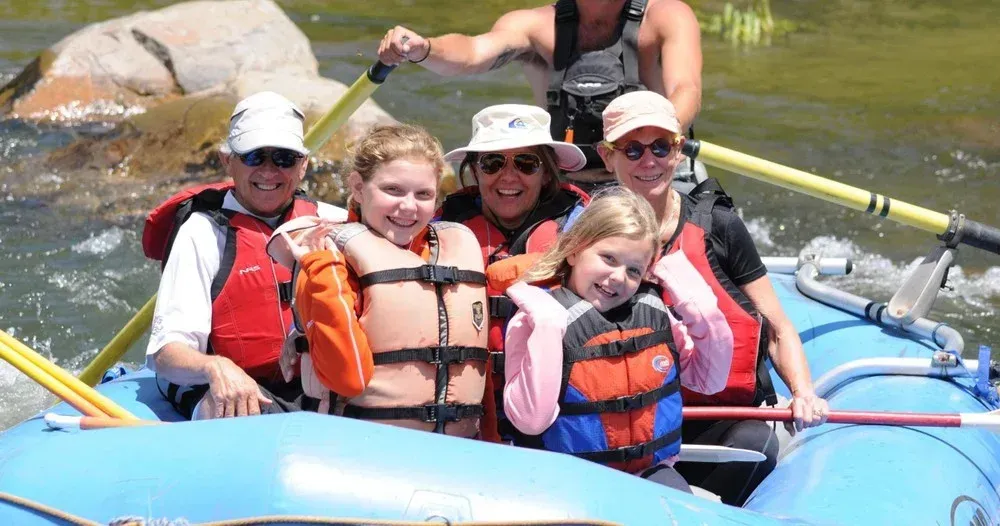 People rafting on a river, smiling, wearing life vests. Blue raft, sunny day.